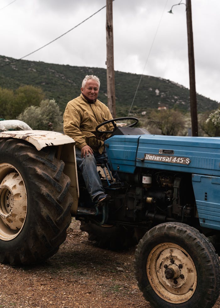 Senior man with gray hair sitting on a blue tractor in a rural landscape.